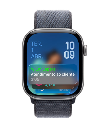 Apple Watch com caixa preta brilhante e pulseira sálvia, com mostrador que exibe uma notificação da Assistência de Espera na parte inferior, a data no canto superior esquerdo e os números das horas organizados no canto superior direito.