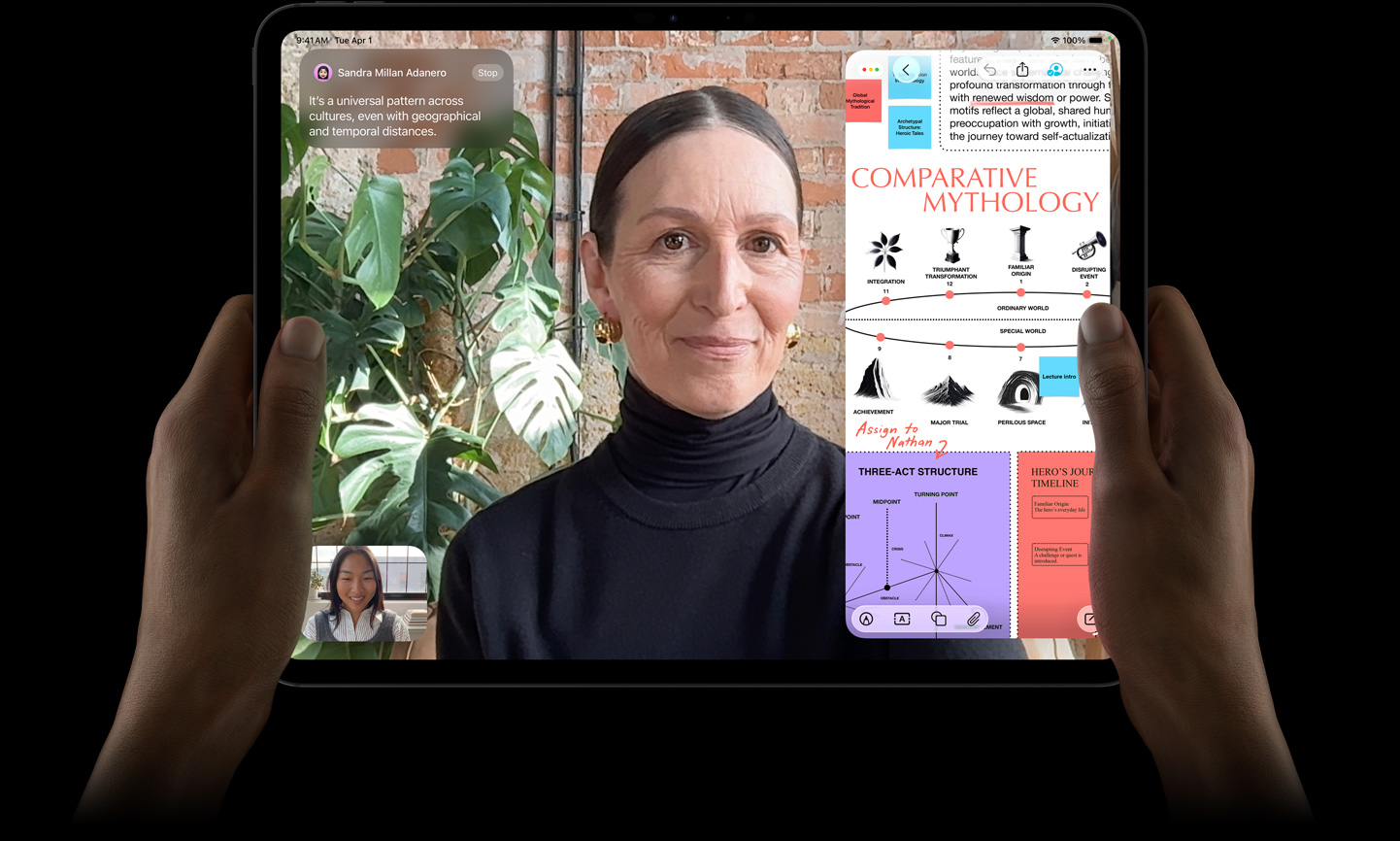 iPad Pro, pohled zepředu, vesmírně černá barva, levá a pravá ruka drží zařízení v orientaci na šířku, zaoblené rohy, černý rámeček displeje, na displeji se zobrazuje konverzace přes FaceTime překládaná v reálném čase a vzdělávací aplikace, ve které je zobrazená prezentace „Komparativní mytologie“ tvořená textem, schématy a ručně psanými poznámkami