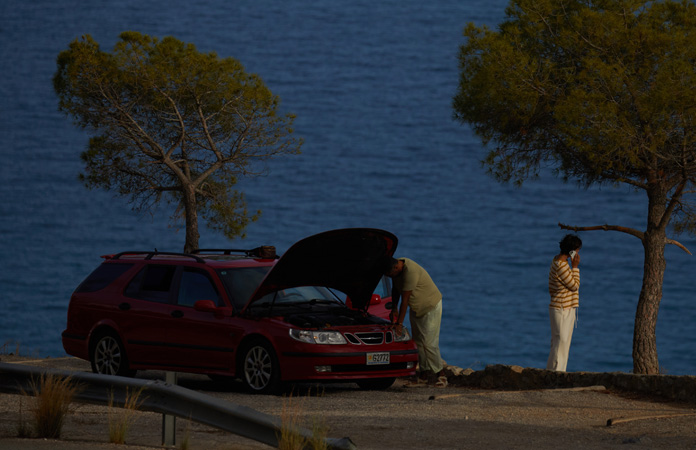 A car stranded by the side of a road, with one person by the car and one person using iPhone to contact roadside assistance