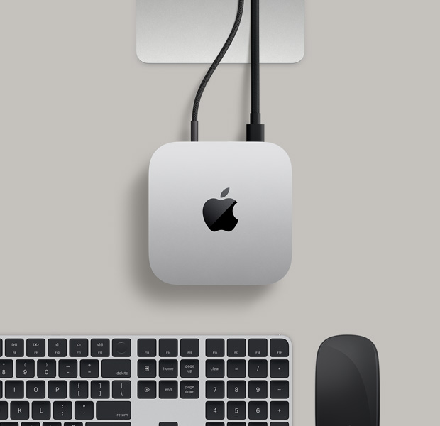 Imagem de cima do Mac mini prateado, com dois cabos conectados nas portas traseiras para um monitor, ao lado de um Magic Mouse preto e um Magic Keyboard preto